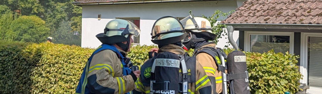 Drei Feuerwehrleute in kompletter Schutzkleidung und mit Atemschutzgeräten stehen vor einem Wohnhaus. Vom Dach des Hauses steigt dichter, heller Rauch auf. Ein roter Feuerwehrschlauch liegt auf dem Gehweg.